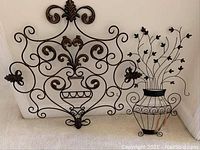 Overall view of two ornate metal wall art pieces: one with large square scrollwork and central vase design, the other shaped like a vase with leafy vine stems