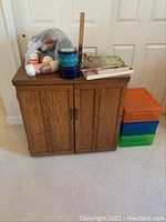 Front view of wooden sewing cabinet with accessories on top including wool yarn, scissors, and storage containers. Colorful plastic boxes next to cabinet.