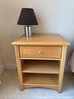 Full view of light wood nightstand with one drawer and two open shelves, with silver base black shade table lamp on top