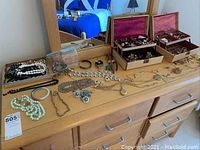 Photo of dresser top with spread assortment of jewelry pieces including necklaces, earrings, pins, brooches, and watches along with two open jewelry boxes.