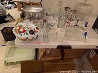 Full view of lot items on table including covered bowl, glassware, silverplate dish, mugs, and roasting pan