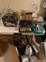 Overview of table with silverplated chafing dish, black case with steak knives, cutlery tray, boxed blender and food processor, and assorted cookware and slow cooker underneath table.