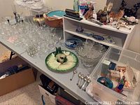 Table covered with clear glassware including wine glasses and punch cups, various ceramic animal figurines, decorative spoons, and a large ceramic chafing dish on a stand.