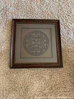 Square framed wall art with circular metal ornamental design in center under glass, ornate frame with dark finish.
