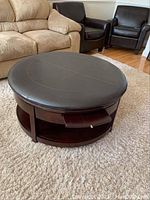 Round padded coffee table ottoman showing full top and base with open shelf and two drawers.