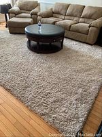 Full coverage photo showing the rug placed in a living room area with beige sofa set and round ottoman on top. The rug is rectangular, 9x12 feet, with plush shag texture.