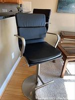 Front angled view of one bar stool showing black cushioned sections on seat and back with metal arms and pedestal base with footrest.