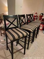 Full view of four black wooden bar chairs with striped beige and black padded seats in a room with carpeted floor and holiday decor in background.