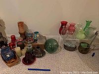 Display of assorted candles and glass vases on carpeted floor with variety of shapes and colors including red, pink, green, blue, and clear glass items.