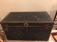 Front view of the vintage black steamer trunk showcasing surface scratches, metal latch closures, and a central lock mechanism.