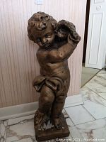 Front view of the painted concrete cherub statue showing detailed facial features, flowing curly hair, and cloth draped around waist holding an object near shoulder.