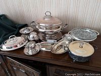 Full view of eleven-piece silver plated serving set with tureen, tray, covered dishes, ladle, and divided dish arranged on wooden sideboard.