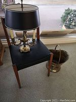 Photo showing mid century wooden chair with black leather-like upholstery, metal table lamp with black shade sitting on the chair, and brass-colored coal bucket next to the chair on the floor.