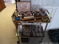 Full view of metal frame bar cart with two wood shelves displaying glassware and bar accessories on top and lower shelves