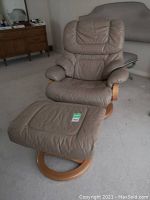 Front view of mushroom-colored leather recliner and footstool set on light carpet. Wooden swivel base visible.