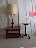 Photo showing a metal base lamp on a wooden drop leaf table and a wooden plant stand beside it against a white wall with closets.