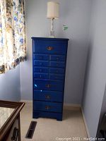 Full view of blue lingerie chest and white ceramic lamp on top next to window with floral curtains.