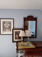 Overview photo showing three framed prints mounted or resting on wall and dressers, two table lamps with cream pleated fabric shades, and a wooden framed mirror with carved detail.