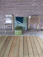 Full view of the lot showing three stacked beige metal folding chairs, a rolled item, vintage card table, small olive green vinyl hassock, and two wooden TV trays with folding legs.