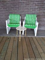 Two patio chairs with green striped cushions and small round table in front of brick wall on wooden deck.