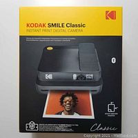 Front image of Kodak Smile Classic camera box showing camera printing a photo. Box is yellow and black with product details and image of camera.