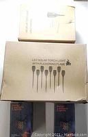 Boxes of LED solar torch lights with flickering flame and light control text; two black boxed solar lantern lights at bottom of pile.