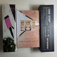 Three boxed items: Pink triple barrel hair waver, tri-fold table makeup mirror, and One Step hair dryer and styler in black box.