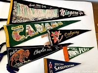 Photo showing full set of 7 felt vintage Canadian pennants including Niagara Falls, Truro N.S., Calgary Alta., Buffalo Chaplin Sask., Percé Rock, and Regina Saskatchewan.