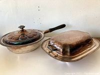 Two vintage silver plated covered dishes, one round with wooden handle and floral lid, one rectangular with side handles and lid, both showing signs of wear and tarnish.