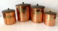 Four vintage copper canisters with brass labels and lids arranged in a line showing different sizes.