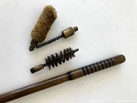 Three cleaning attachments lying next to the wooden rod: wool mop brush, twisted wire brush with bristles, and cylindrical bristle brush with brass fittings.