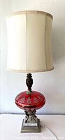 Full view of the vintage red glass table lamp including the white fabric lampshade, ornate red glass base with silver decorative floral detailing and marble base.