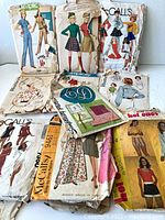 Photo showing 11 vintage sewing pattern envelopes with illustrated designs for women's and girl's clothing including skirts, dresses, tops, and shorts. Includes brands Simplicity, McCall's, Butterick. Patterns appear worn with folded, torn edges.
