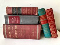 Full view of all five vintage hard cover dictionaries and reference books stacked horizontally and vertically against a white wall.