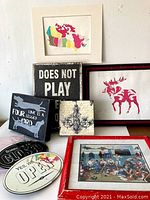 Photo showing colorful framed map of Canada, wooden signs with various texts including 'DOES NOT PLAY' and 'LOVE IS A FOUR LEGGED WORD', chandelier illustration plaque, framed red moose silhouette, and a framed hockey group photo with red border.