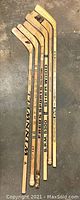 Six wooden hockey sticks displayed side by side on a concrete floor, showing full length including shafts and blades, branded with names like Sher-Wood and Winwell.