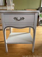 Front view of white night table showing drawer with ornate handle and lower shelf.