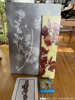 Grey canvas with white floral silhouettes next to tall orchid panel and small block and matted picture with cartoon and French motifs seen on floor
