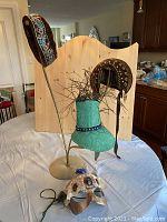 Brass hat stand displaying two brown velvet hats and a green fascinator with peacock feathers, with a decorated theatre mask in front on the table.