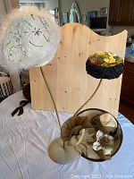 Photo showing three hats displayed on a hat stand: a white/beige hand-beaded beret with feathers, a black felt hat with colorful rosettes, and a beige fascinator-style felt hat with fabric flowers.