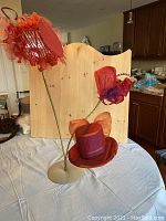 Three red dramatic hats displayed on a metal stand: one large rounded mini hat with red/orange striped fabric and orange mesh ribbon, with two smaller fascinators made of red netting and purple accents on long thin stems.