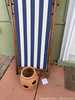 Wood-framed lounge chair with blue and white striped fabric leaning against a green wall with a terracotta strawberry pot placed on the ground in front. Chair folds flat with visible slight wood nick.