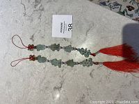 Two Asian jade decorative hanging ornaments on a marble surface with red tassels and red string loops, showing condition and overall design.
