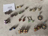 Overview of 16 pairs of costume earrings featuring floral, beaded, and enamel designs on French wire hooks and a few clip-ons, shown on a beige surface.