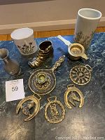 Photo shows three vases - one porcelain with purple floral design, a metal pewter vase with crown emblem, and another plain metal vase; a solid brass boot-shaped figurine; a brass ornate candle holder with handle, and multiple brass appliques including horseshoes and horses, as well as a brass tea strainer.