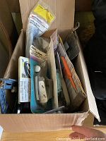 Photo showing box with assorted hardware parts and tools, including electrical outlet, extension cord, mounting bracket, wire clamp, and garden trowel