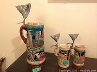 Large and small German beer steins with decorative relief designs and pewter lids displayed on a surface, showing size comparison and intricate craftsmanship.