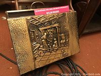 Front view of a rectangular brass magazine holder with a hammered texture and a raised relief showing a scene with two people by a fireplace.