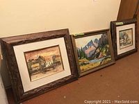 Three framed original paintings leaning against a wall, showing different scenic subjects, all framed in carved wooden frames with beige mattes