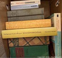 Photo of various cookbooks and specialty books in a box, showing titles like Betty Crocker's Cookbook and General Foods Kitchens Cookbook.